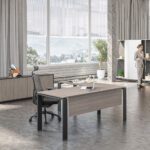 Modern L-shaped office desk with a light wood grain top and black metal legs in a spacious, well-lit office setting with a large window view.