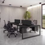 Modern, industrial-style office workstation featuring a long black desk supporting four seating positions separated by low black privacy screens. Two black mesh ergonomic chairs are visible, one with a laptop, set against a background of rough concrete walls and large windows.