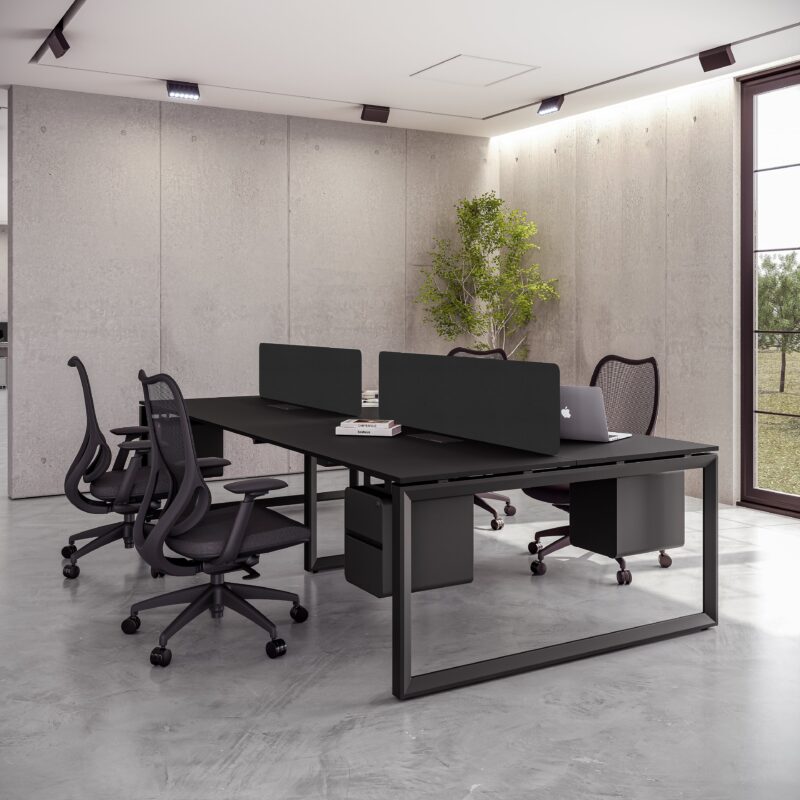 Modern, industrial-style office workstation featuring a long black desk supporting four seating positions separated by low black privacy screens. Two black mesh ergonomic chairs are visible, one with a laptop, set against a background of rough concrete walls and large windows.