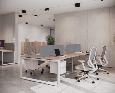 Spacious and bright modern office workstation featuring a long light wood desk with white legs, offering multiple seating areas separated by gray privacy panels.