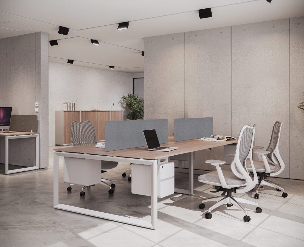 Spacious and bright modern office workstation featuring a long light wood desk with white legs, offering multiple seating areas separated by gray privacy panels. Several white mesh ergonomic chairs are positioned around the desk, and a laptop is open on the surface. The background shows concrete walls and other office furniture.