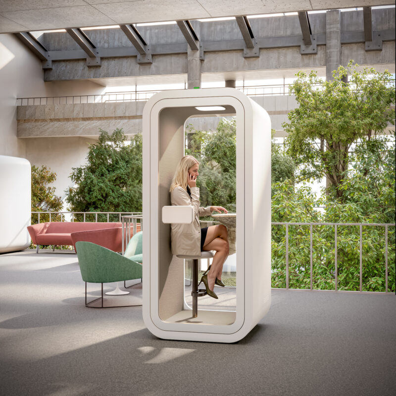 Woman taking a phone call inside a modern, soundproof white office pod with a built-in counter/desk, situated in a large, bright open-plan office area with abundant greenery.