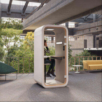 Single-person soundproof meeting pods with rounded corners, featuring a woman taking a phone call while seated at a small desk inside a bright, multi-story office atrium with surrounding greenery.
