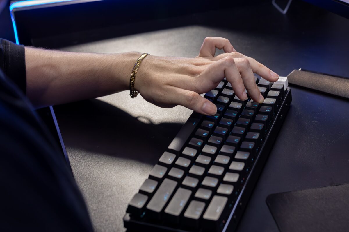 A close-up shot of a person's left hand wearing a gold chain bracelet, typing on a mechanical gaming keyboard with dark gray keycaps and subtle blue backlighting.