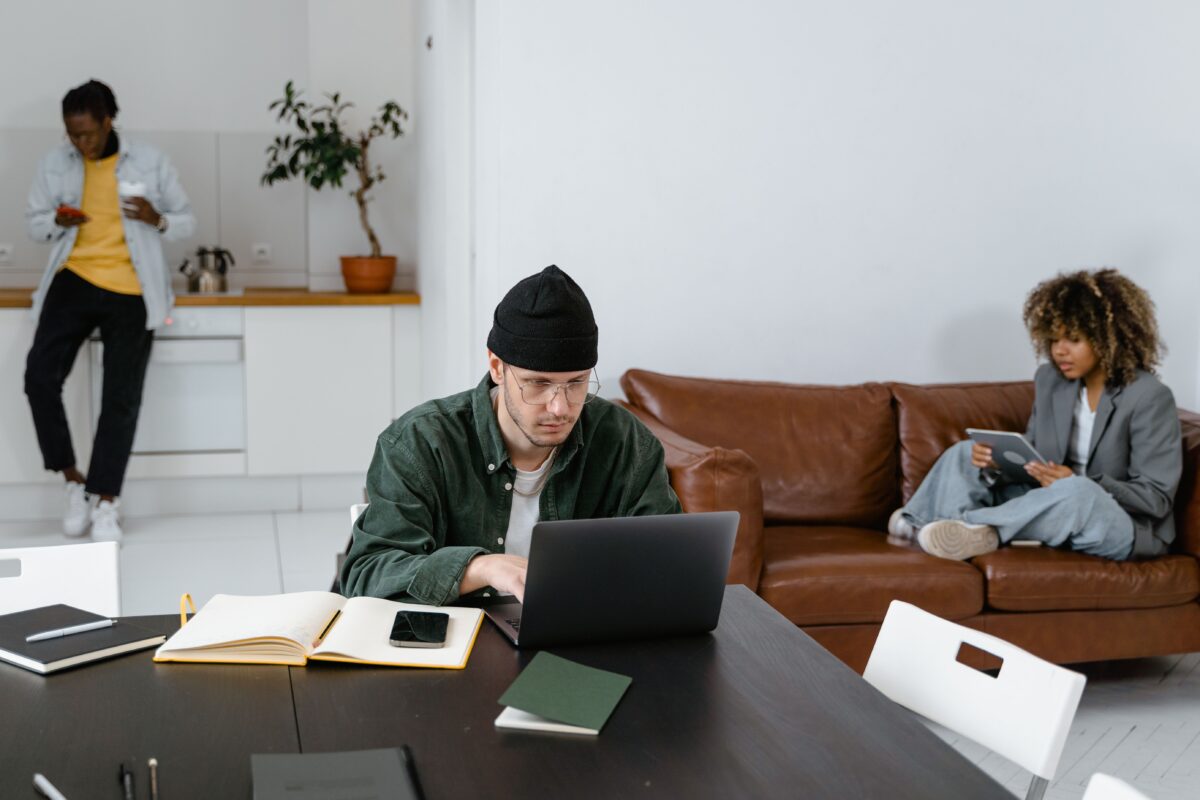 Three people working independently in a modern co-working space. A man works on a laptop at a table in the foreground, a woman uses a tablet on a brown leather sofa, and another man looks at his phone while standing in a kitchen area.
