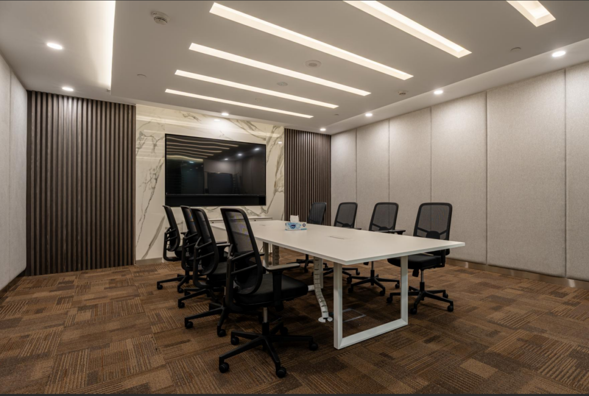 A modern conference room featuring a long white meeting table surrounded by black mesh office chairs, set against a brown patterned carpet and a wall with a large TV display.